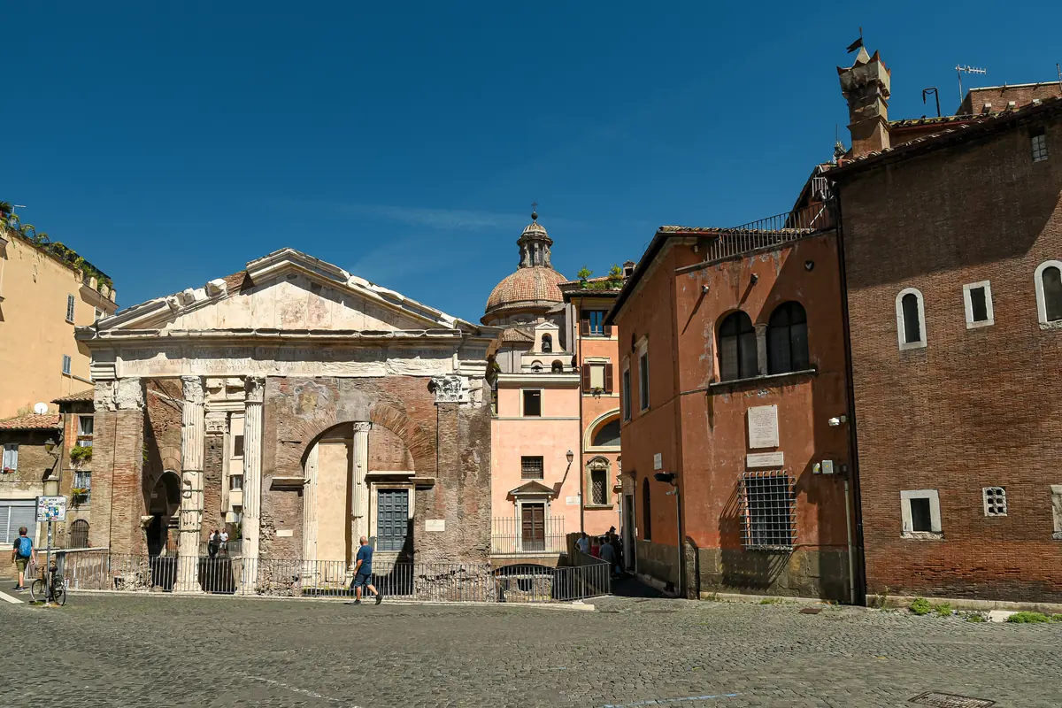 The Portico d'Ottavia in Rome: a pedimented ancient gateway of brick and stone at the edge of a cobbled square, with the church of Sant'Angelo in Pescheria built into it